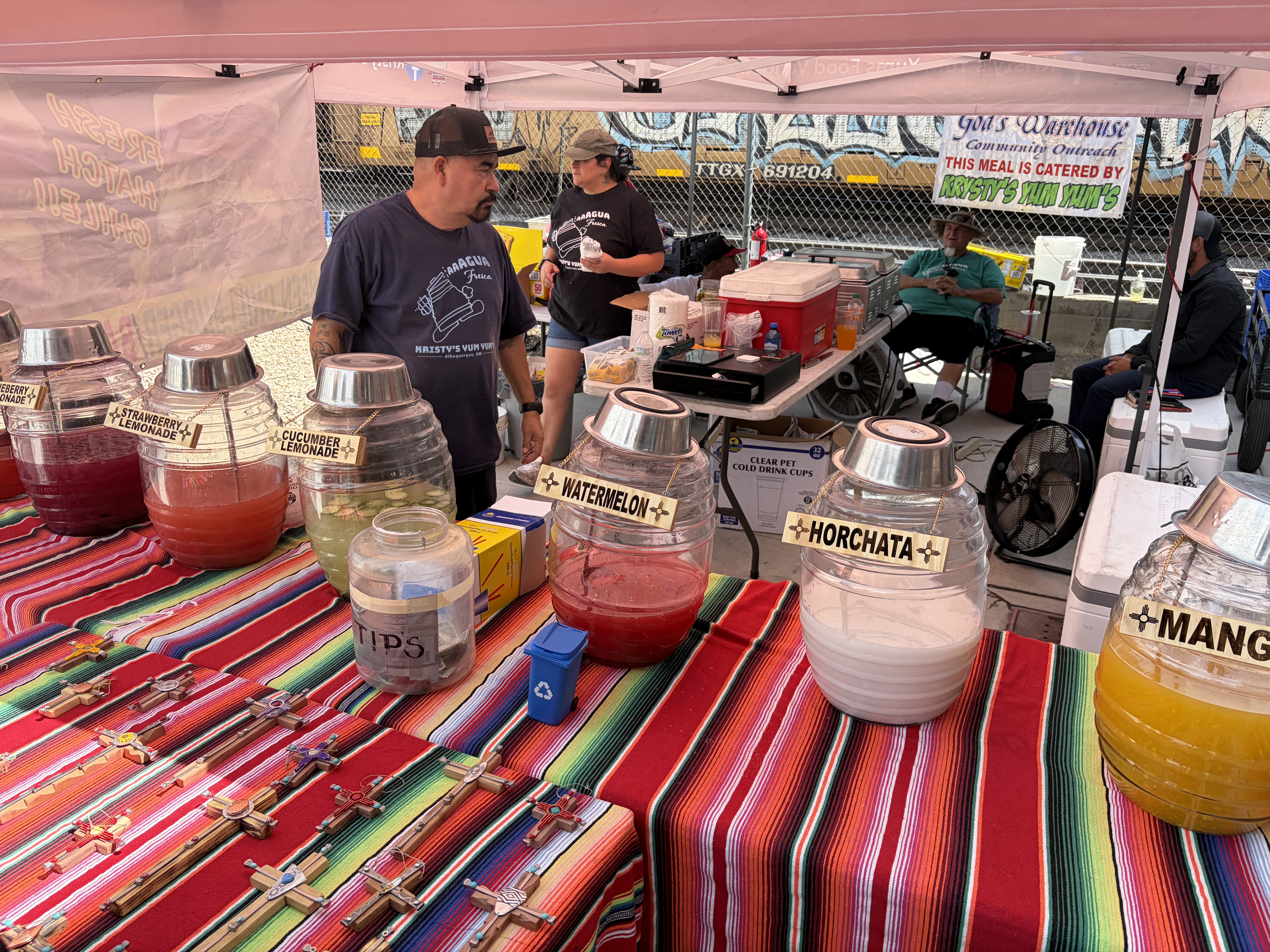 Agua fresca dispensers labelled strawberry lemonade, cucumber lemonade, watermelon, horchata and mango on a serape-covered table, wooden crosses for sale, a railcar behind the fence.