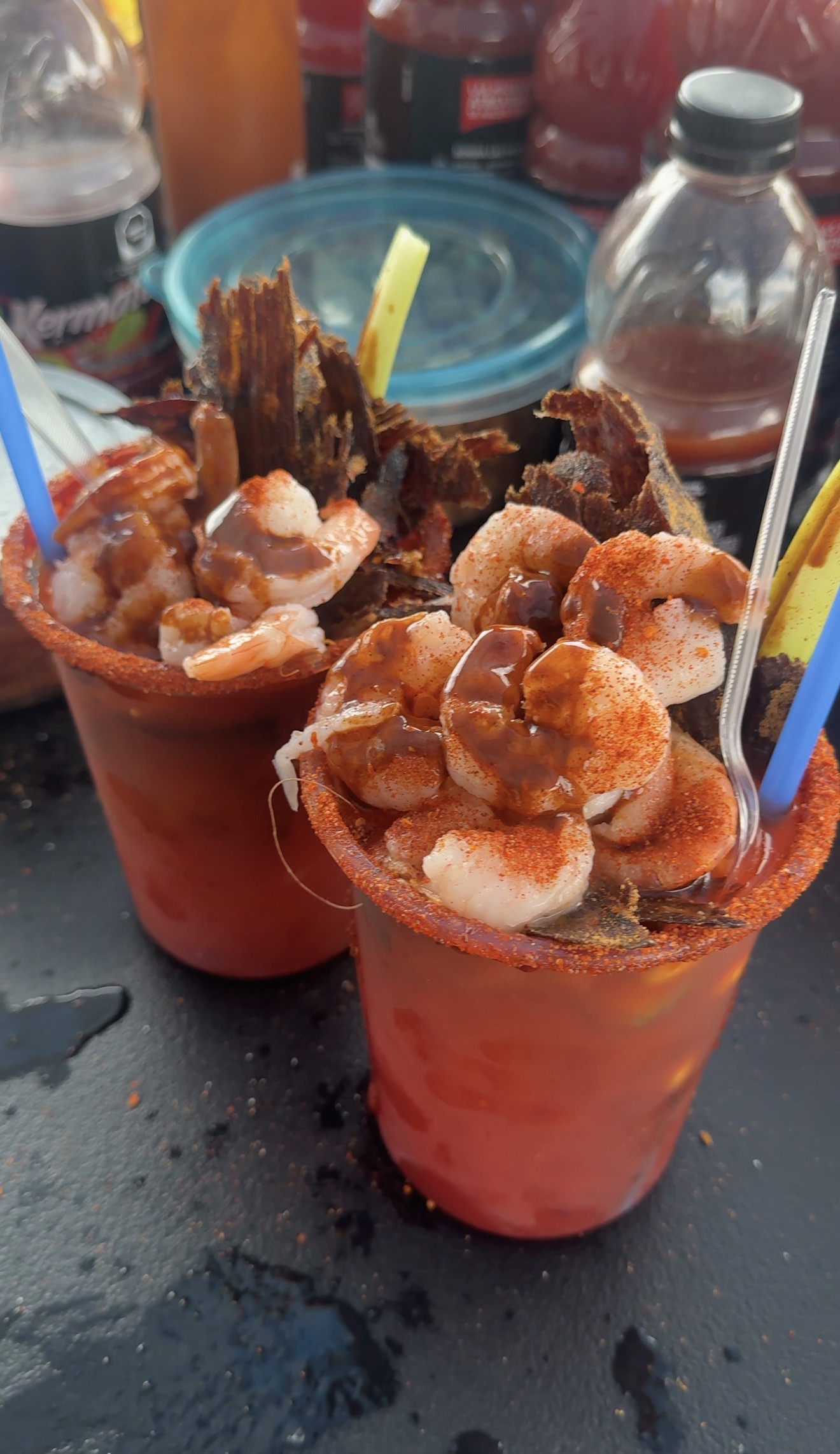 Two chamoy-rimmed cups of red Kermato topped with shrimp, chile powder and standing strips of dried meat, with tamarind straws.
