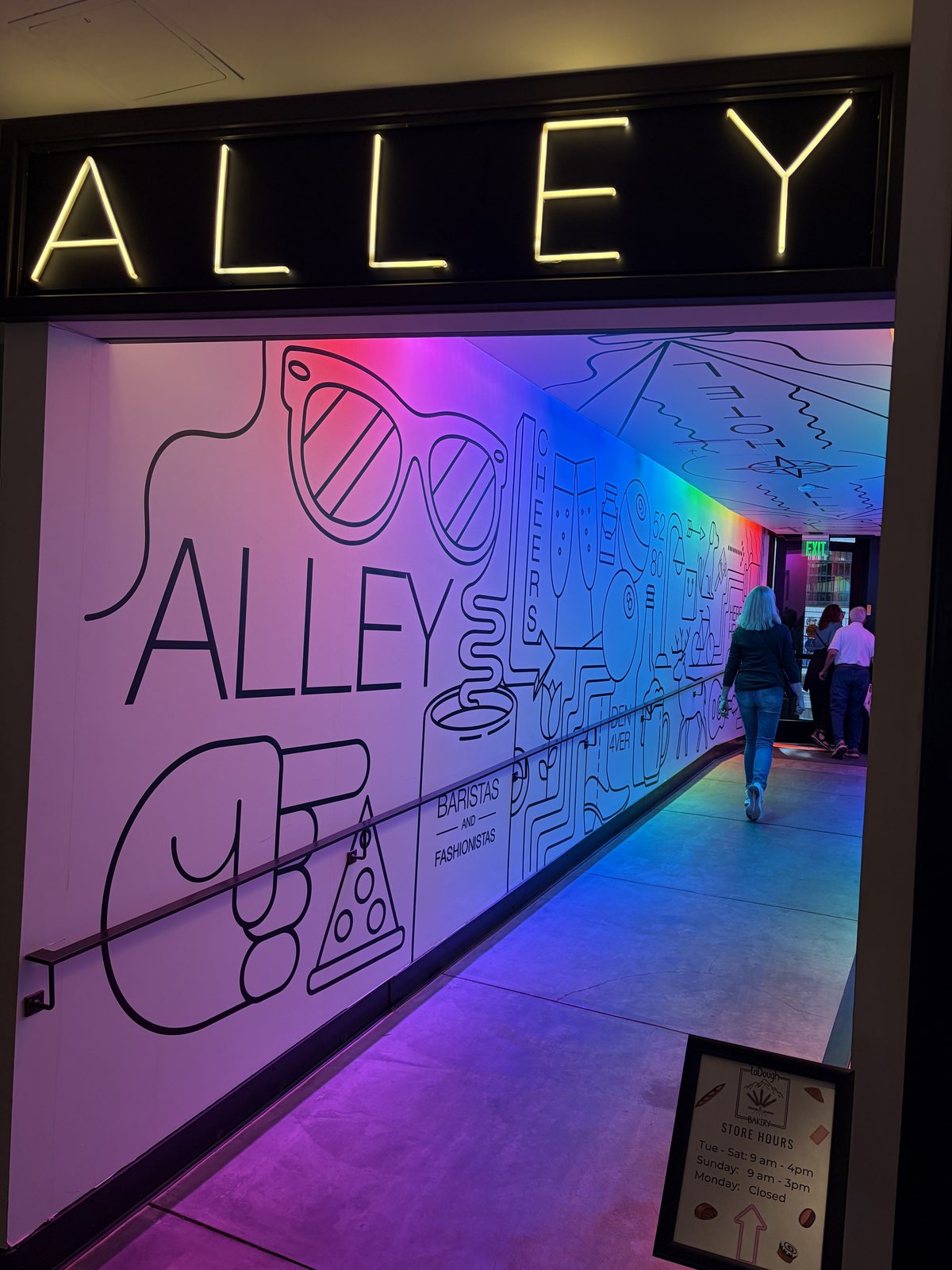 A lit ALLEY sign in neon capitals above a corridor washed in pink and violet light, line-drawn mural along the wall.