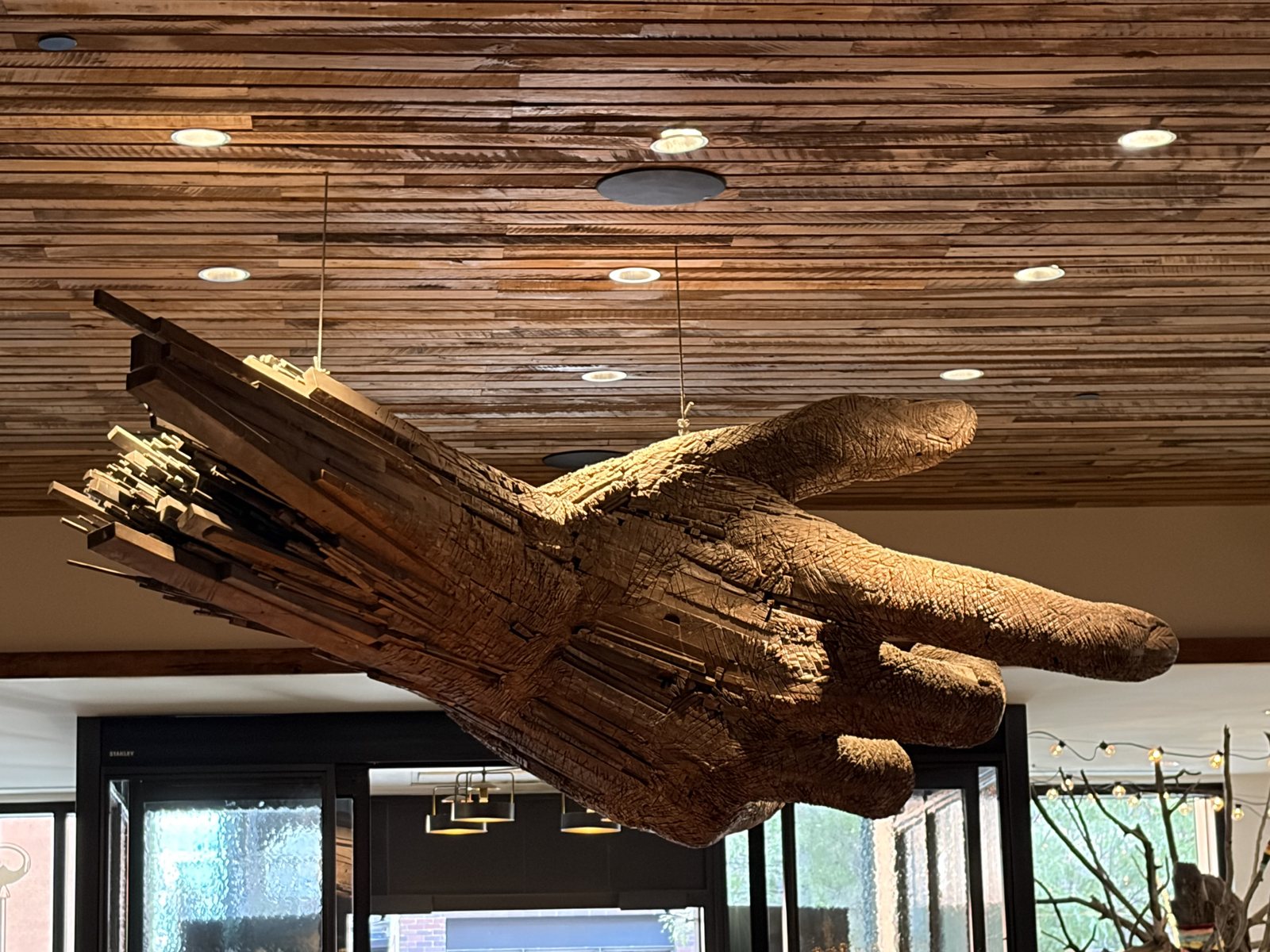 A large carved wooden hand suspended from a reclaimed-wood ceiling, index finger extended, pointing.