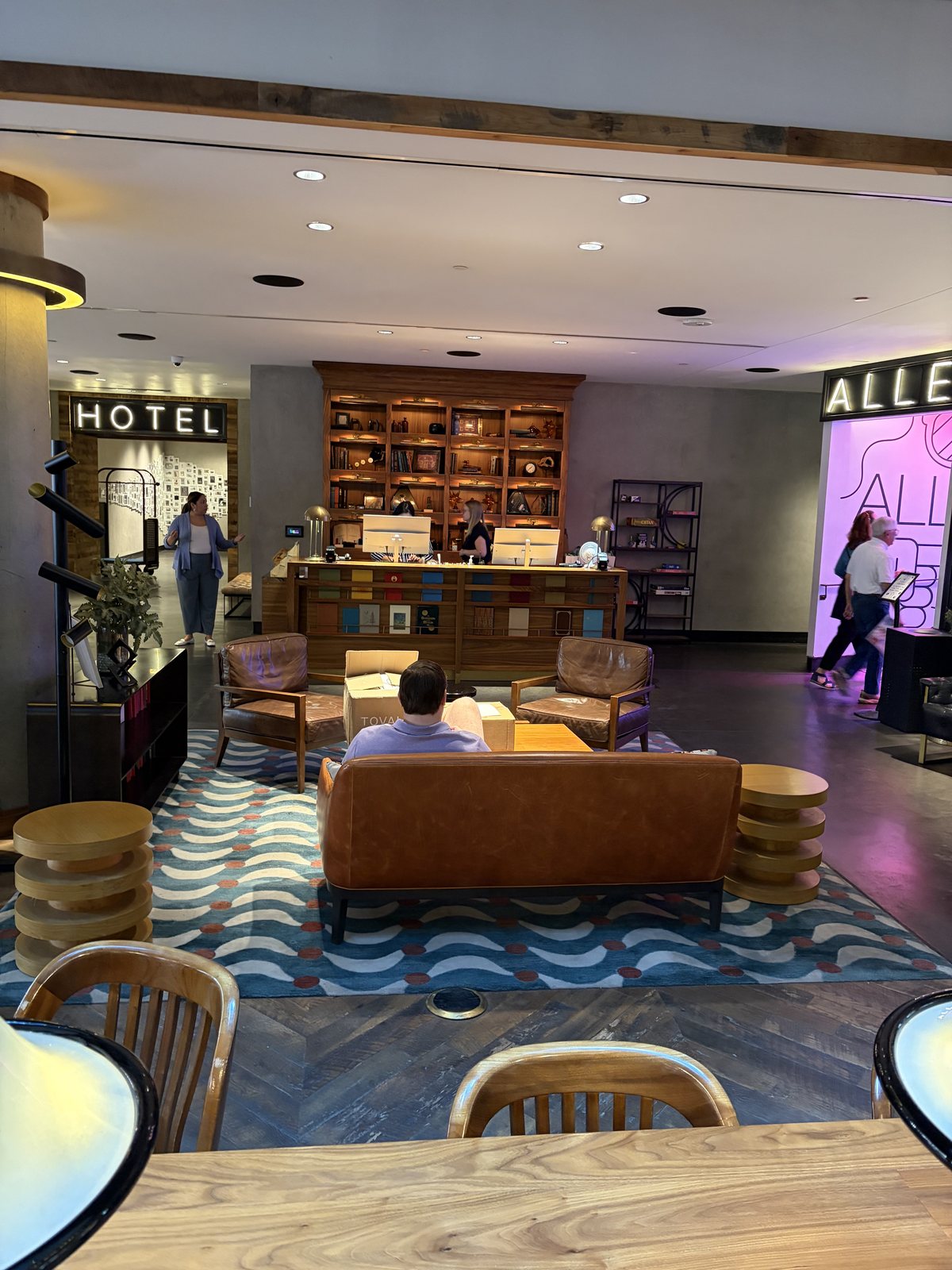 A hotel front desk under a lit HOTEL sign in thin neon capitals, dark wood, low lobby seating.