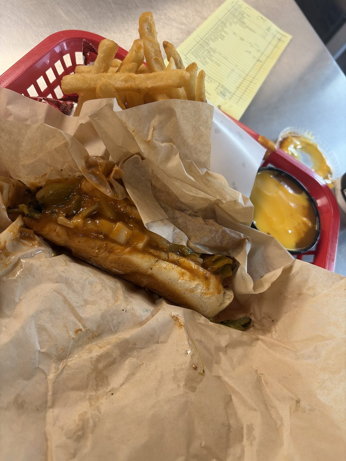 A chili dog smothered in red and green chile, wrapped in two sheets of parchment paper in a red plastic basket, beside french fries and a cup of nacho cheese mixed with red chile.