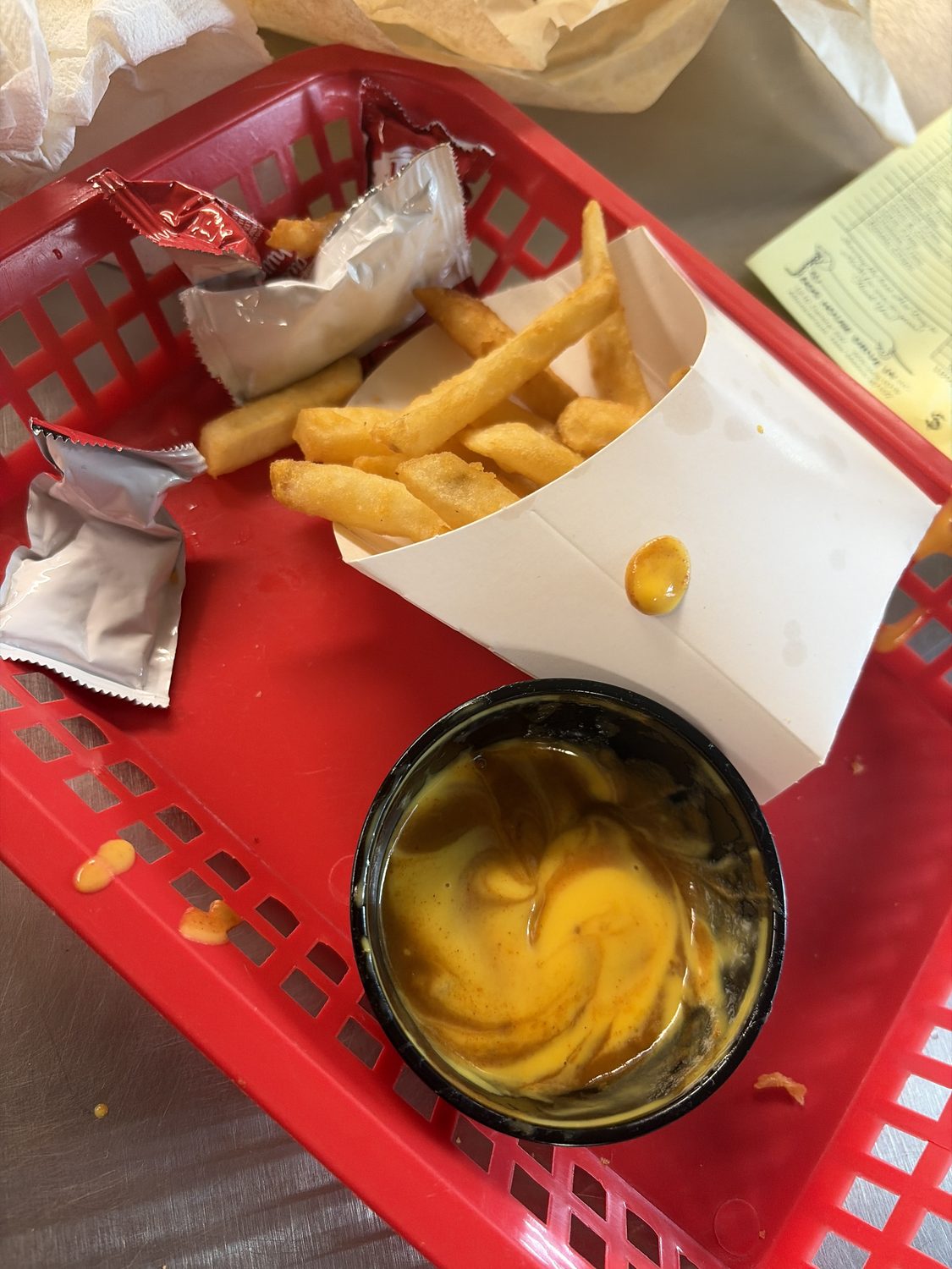 French fries in a red plastic basket beside a paper cup of nacho cheese swirled with red chile, an unopened ketchup packet resting on the tray.