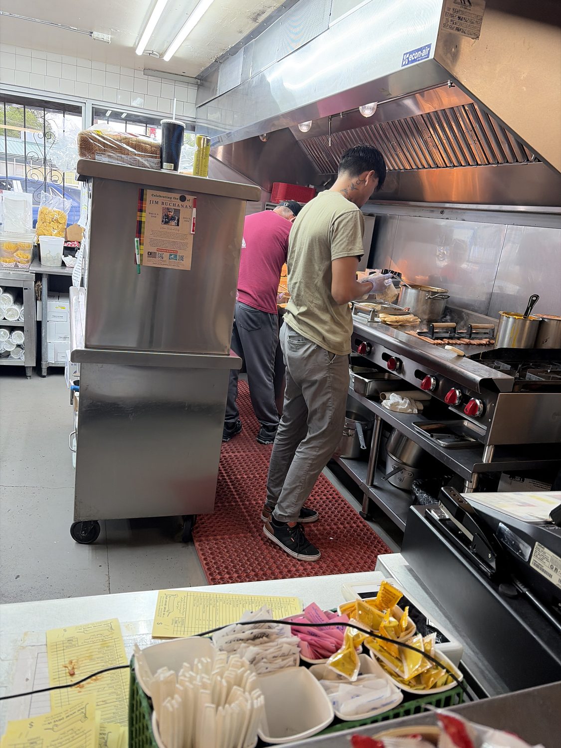 Two cooks working a stainless steel line under a hood, both with their backs to the camera, order tickets and mustard packets stacked in the foreground.