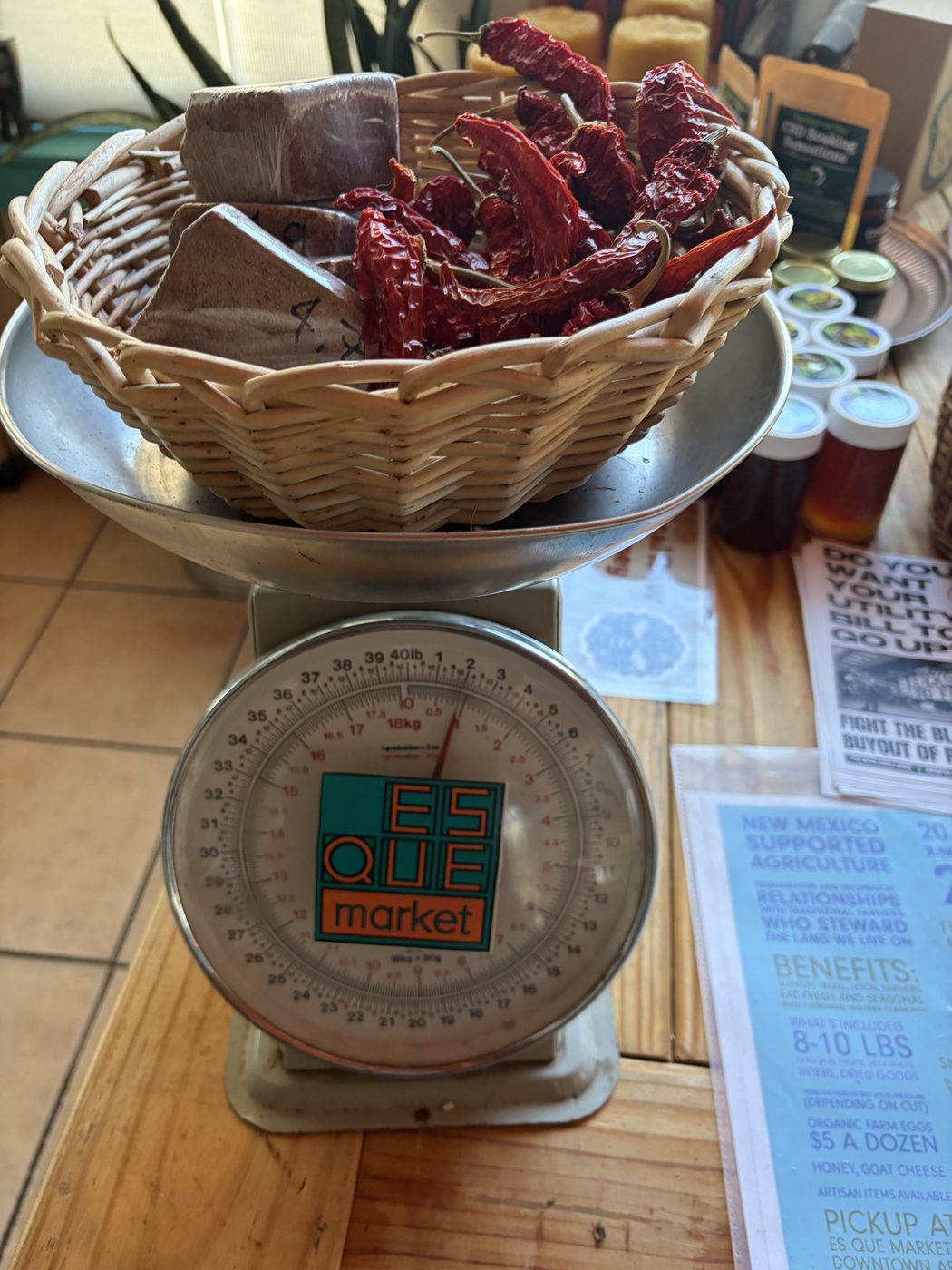 A wire basket of dried red chile pods sitting on a vintage mechanical scale with an ES QUE market dial face.