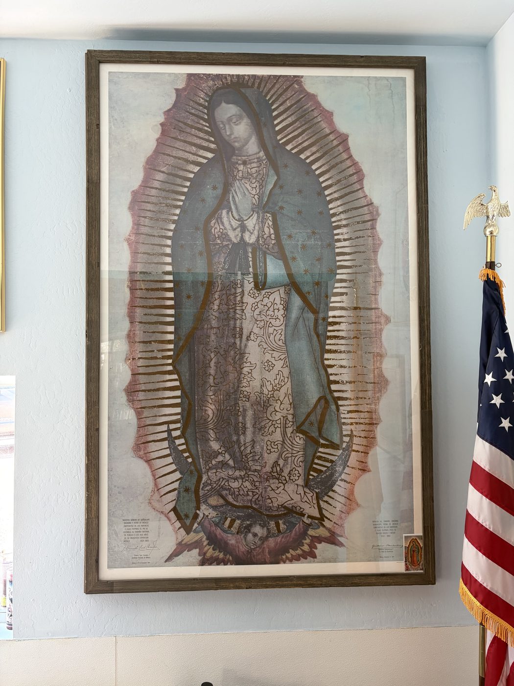 A framed image of the Virgin of Guadalupe on a white wall beside a United States flag.