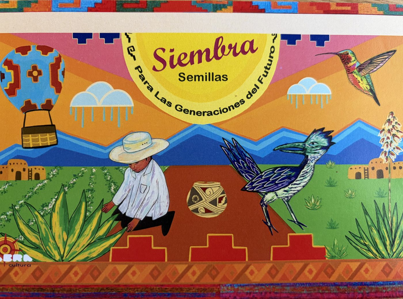 A bright mural: a hot air balloon, a farmer in white with a straw hat sowing seed, a roadrunner, agave and adobe under blue mountains, banner reading Siembra Semillas Para Las Generaciones del Futuro.