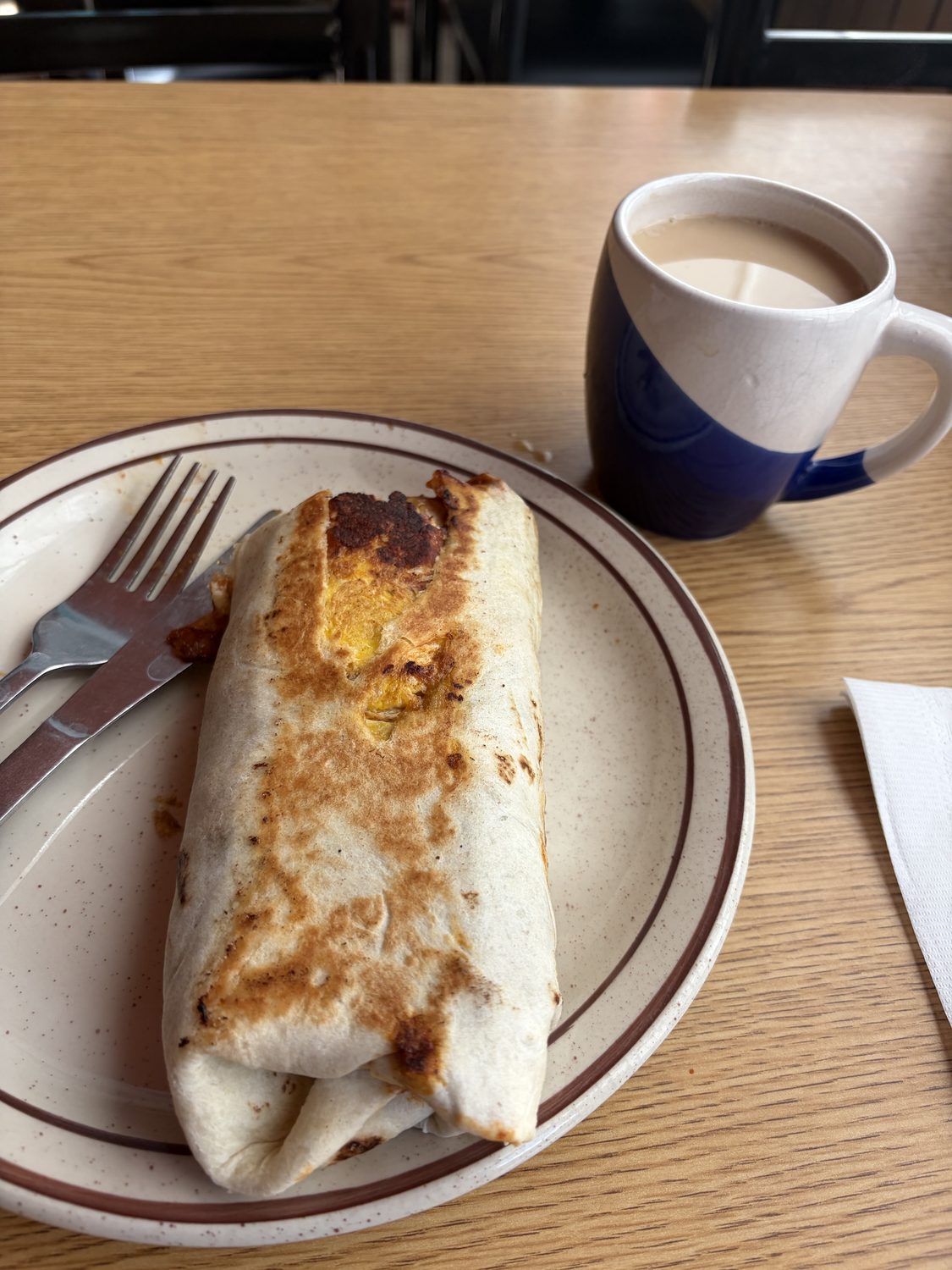 A large burrito on a rimmed plate, lightly toasted golden brown, beside a blue mug of coffee.