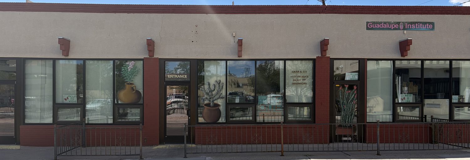 A long stucco and red-brick storefront behind an iron railing. Painted cactus and clay pot murals fill the windows, ES QUE market's logo stands inside the glass, and hand-lettering reads GRAB & GO, FRESH PRODUCE, SALADS AND SANDWICHES.