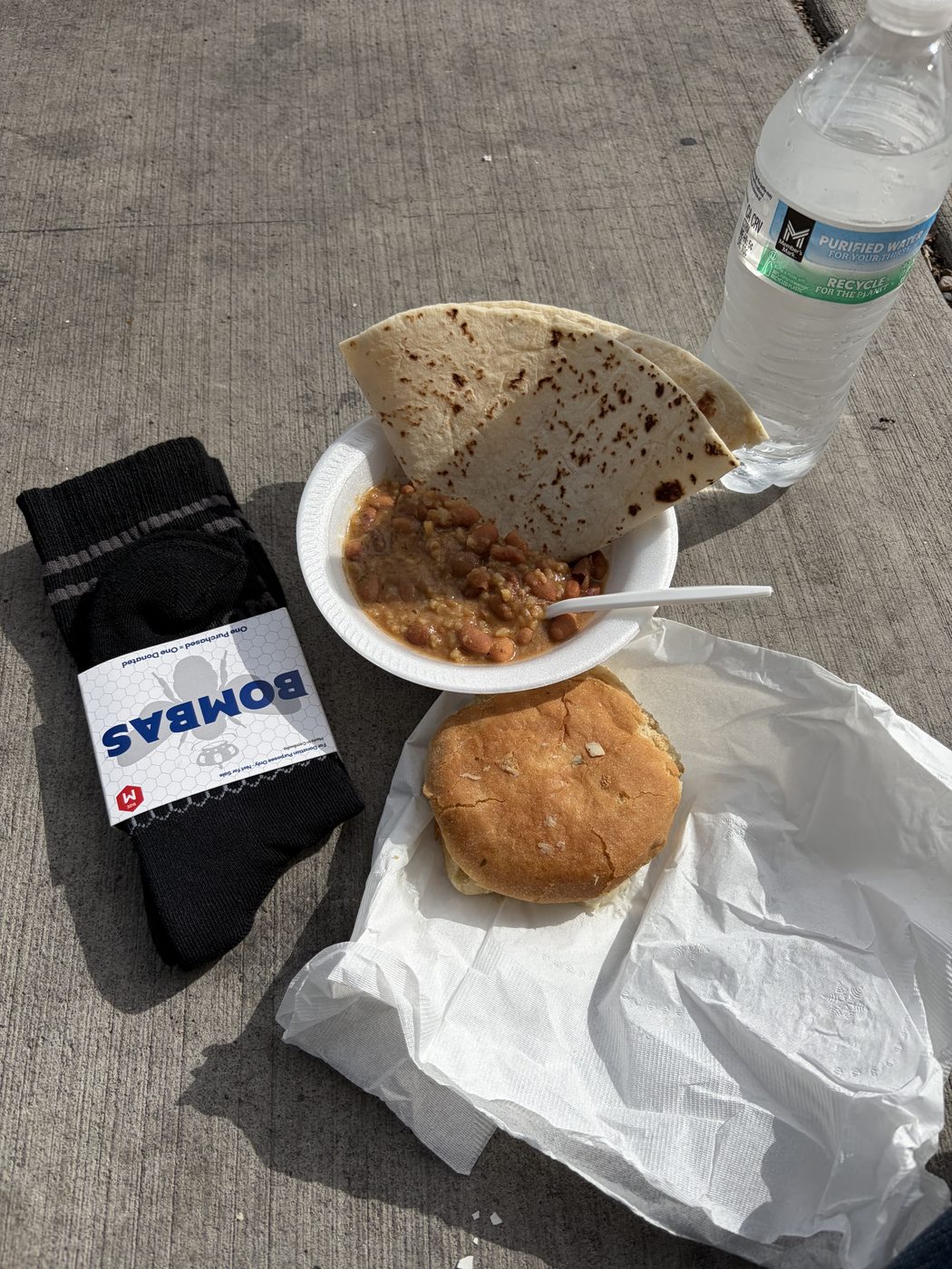 A paper bowl of beans with a folded tortilla, a fried bread, a sealed bottle of water and a new pair of socks, set on the pavement.
