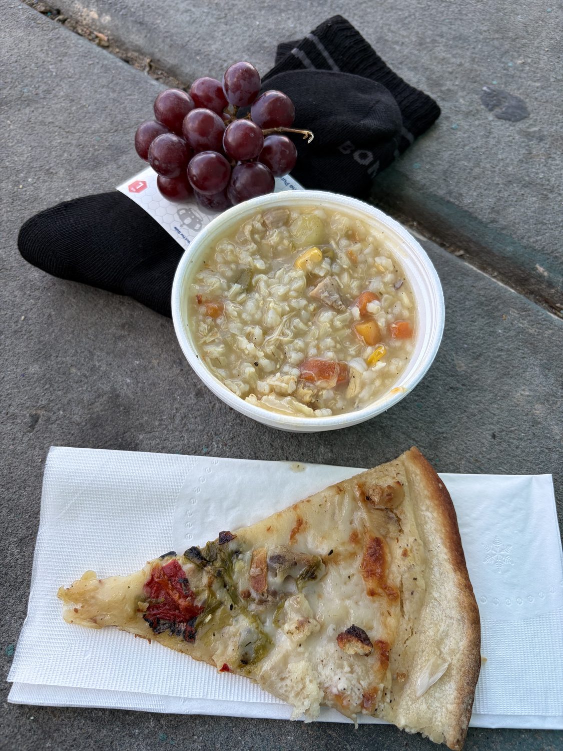 A bowl of chicken and rice soup, a slice of cheese pizza on a napkin, a pair of black socks and a bunch of red grapes, set out on a pavement kerb.