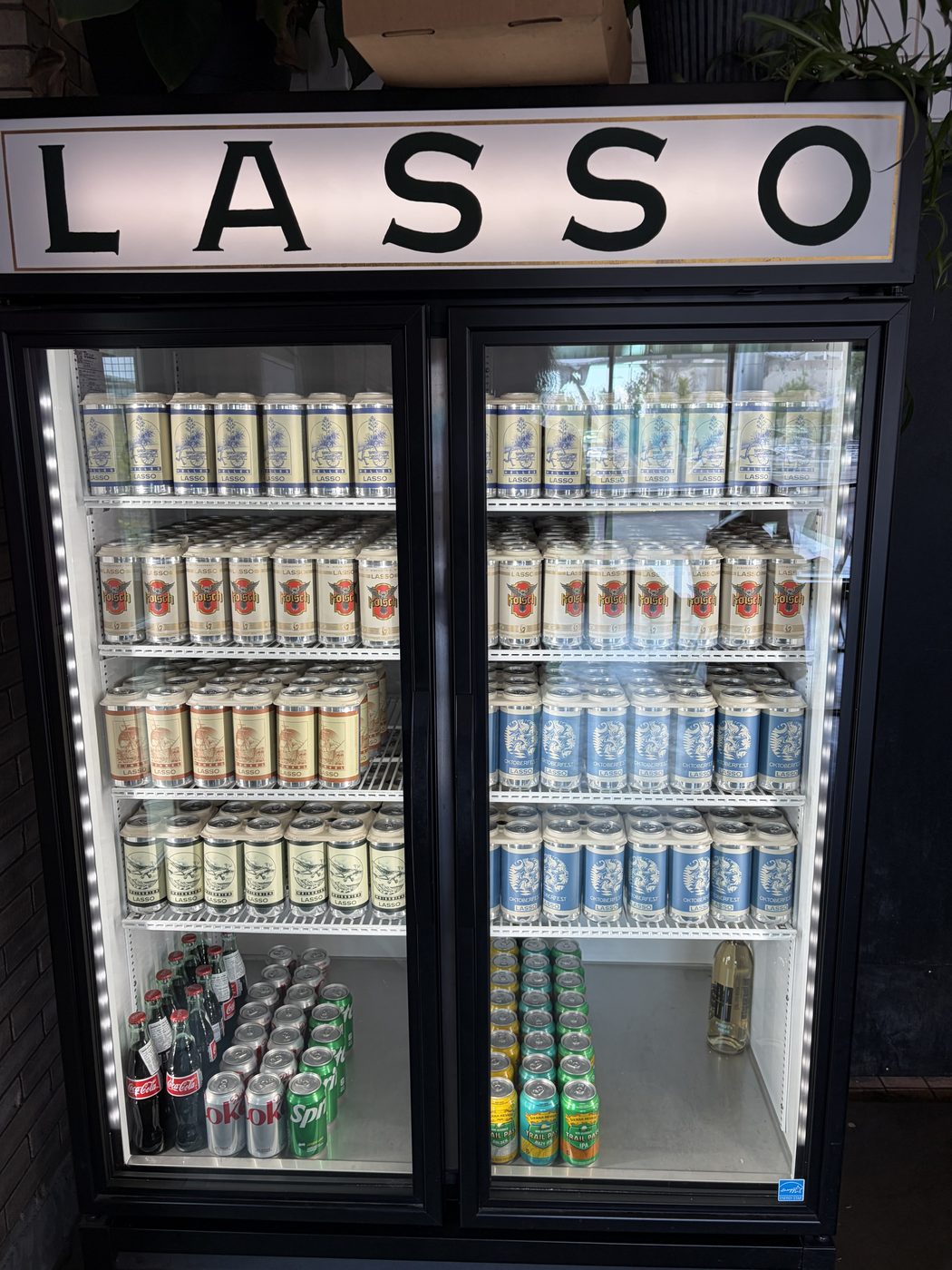 A two-door cooler under a lit LASSO sign, filled with shelves of four-packs: cream and blue Helles, red-crested K&ouml;lsch, rust Dunkel and pale blue Oktoberfest.