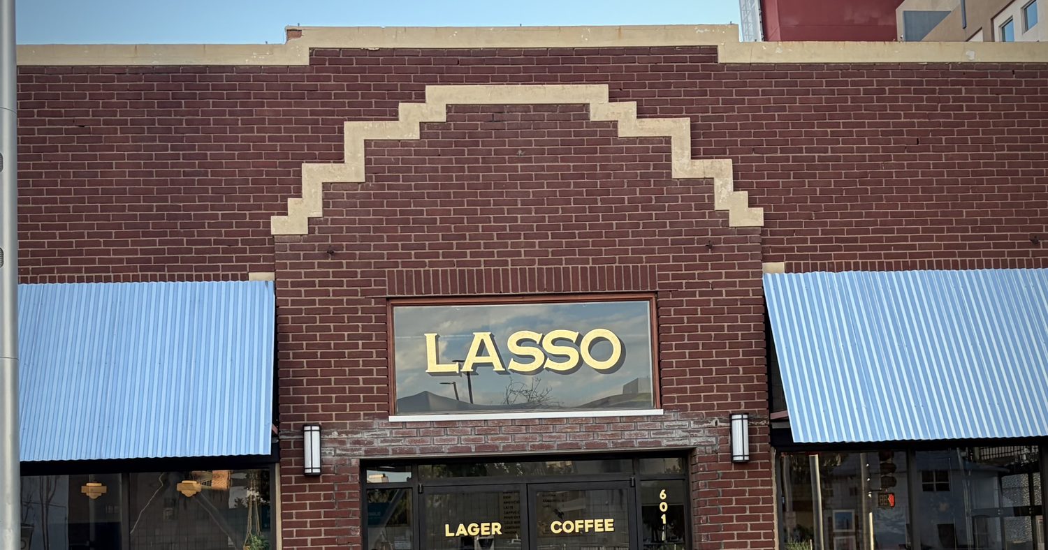 A dark red brick storefront with a stepped parapet, blue corrugated awnings, and a gold LASSO sign on the transom above double glass doors lettered LAGER and COFFEE, with the number 601 in gold beside them.