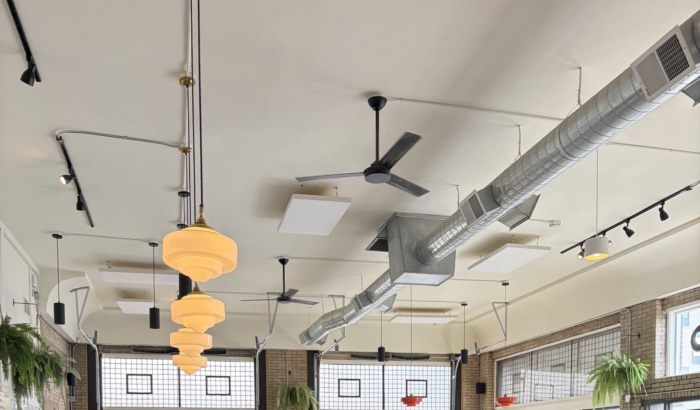 Exposed spiral ductwork and ceiling fans above tall glass-block windows, with amber schoolhouse pendants hanging in a row.