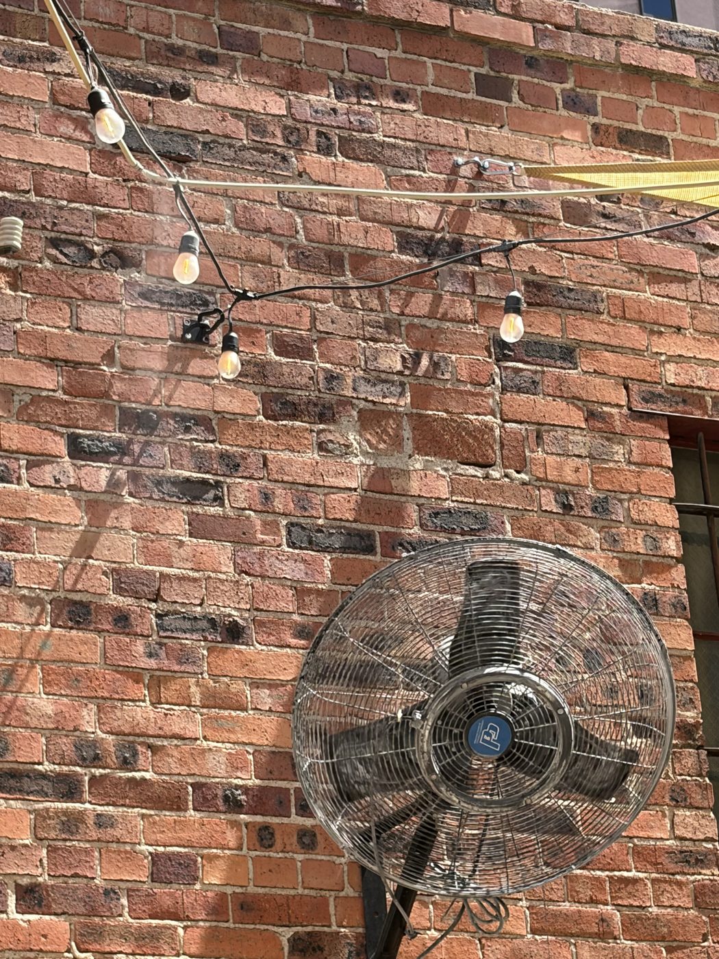 A large industrial fan mounted on a brick wall with string lights strung across it.