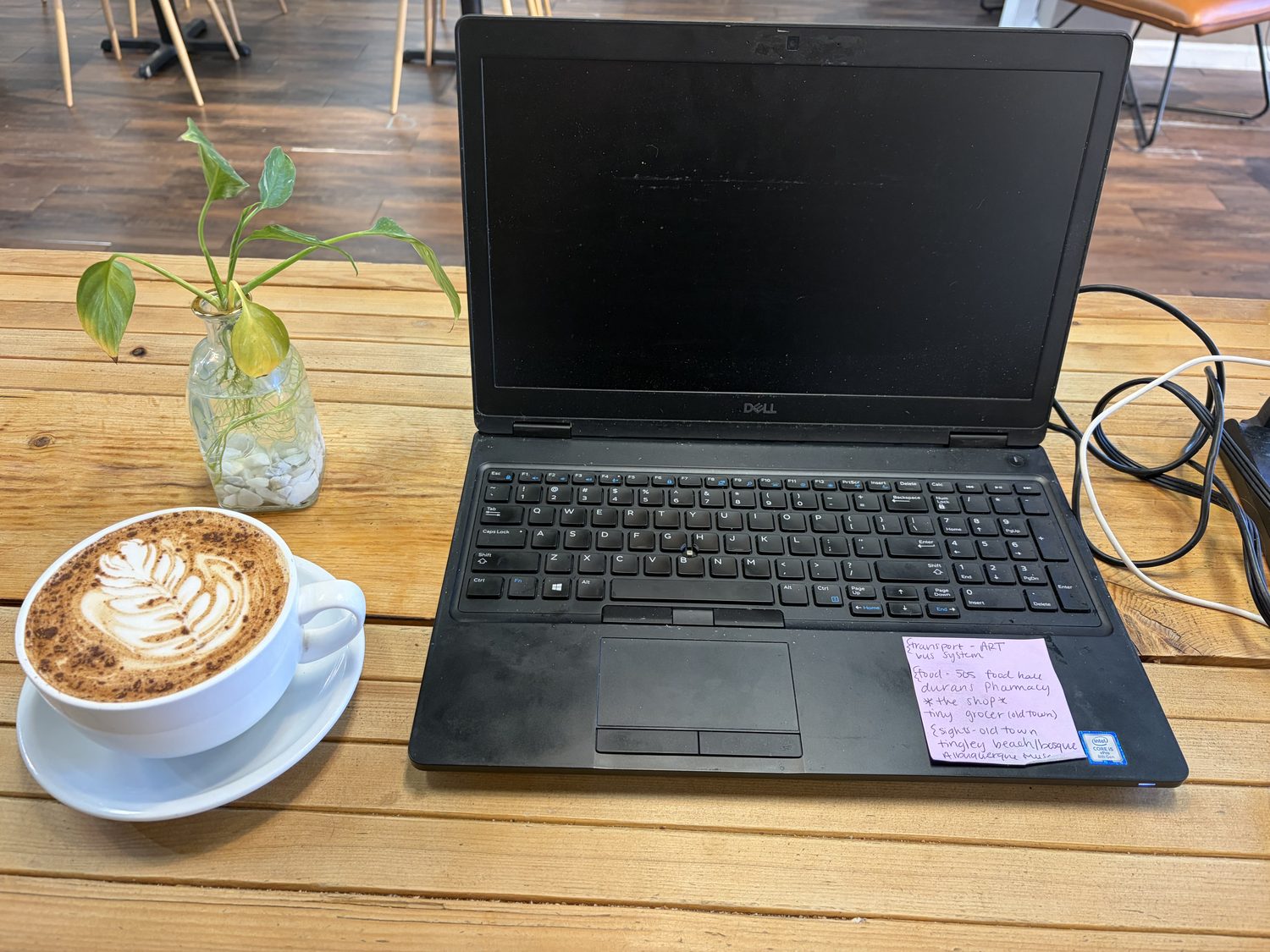 A cappuccino-style latte with a rosetta poured in the foam, sitting on a saucer on a pale wooden table beside an open laptop, a cutting from a plant in a glass jar, and a pink sticky note covered in handwriting.