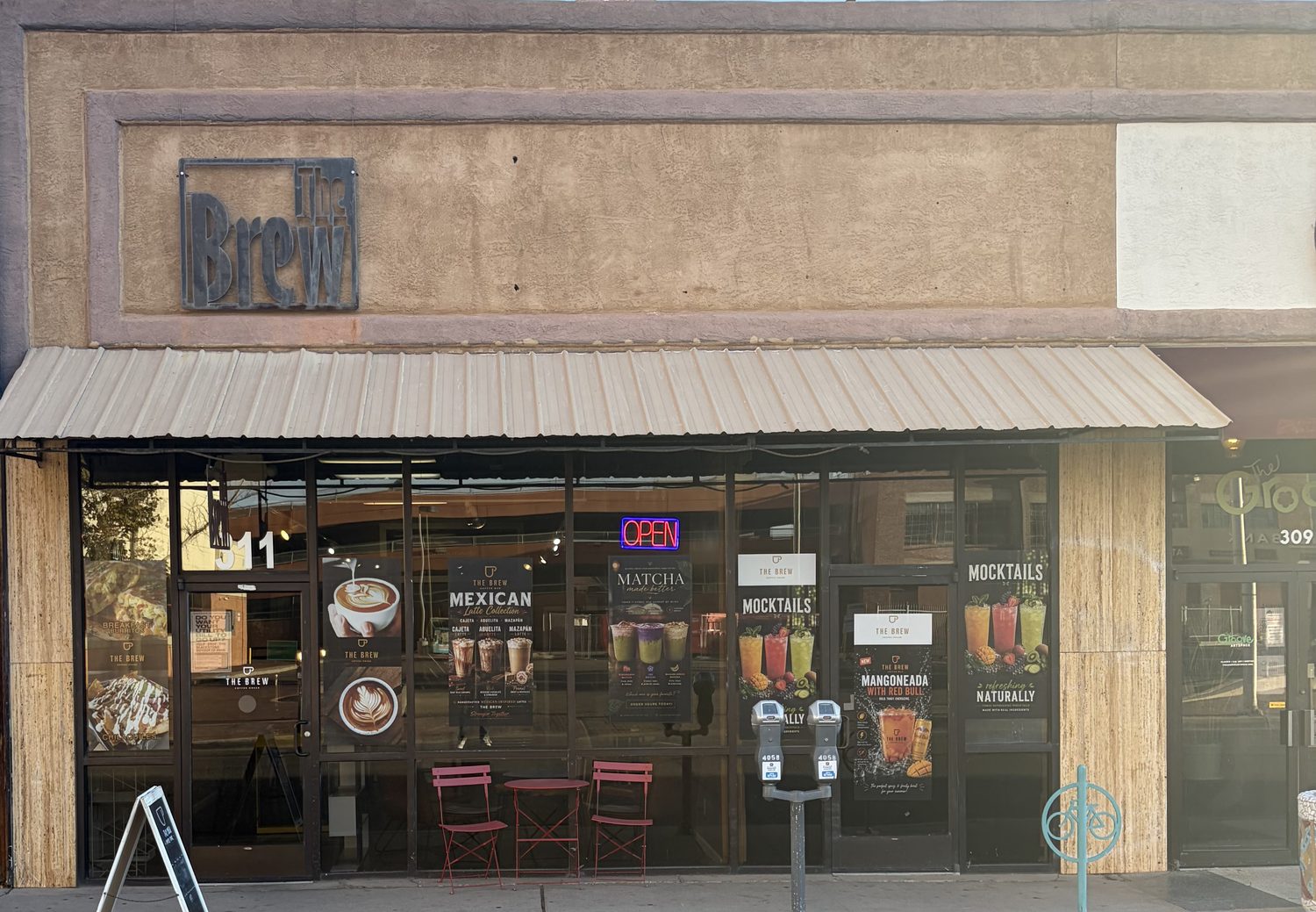A stucco storefront under a metal awning with THE BREW lettered on the wall, an OPEN neon sign, red stools inside, and window posters for a Mexican latte collection, matcha and mocktails.
